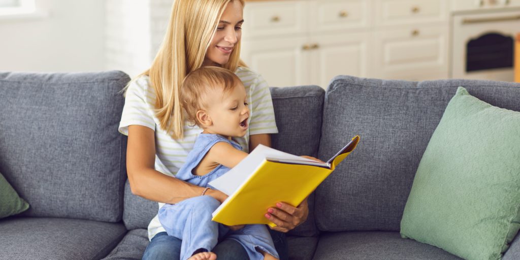 National Social Worker Day. Young woman reading to a toddler sitting in her lap on a gray sofa.