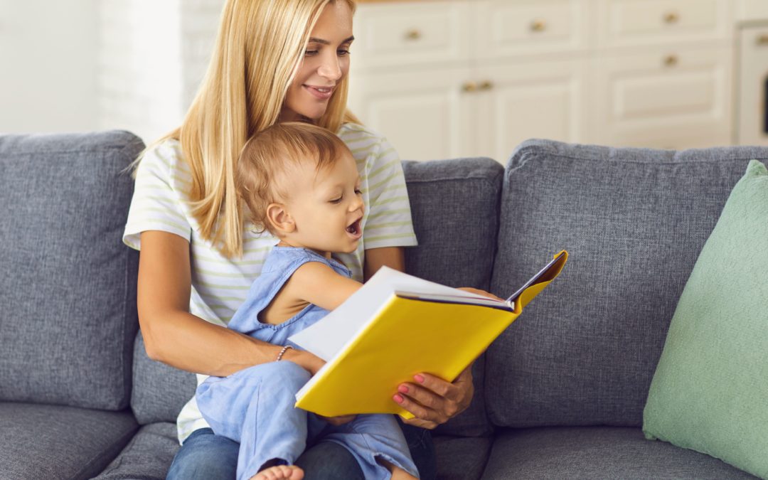 National Social Worker Day. Young woman reading to a toddler sitting in her lap on a gray sofa.