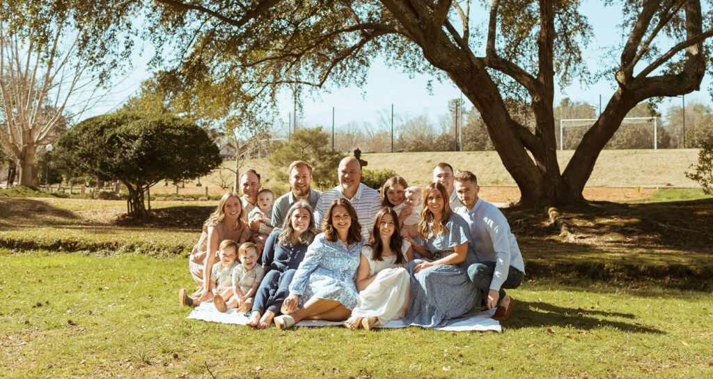 Kim's Story: Kim Trainer's family sitting on a picnic blanket outside on a field of green grass under a tree.